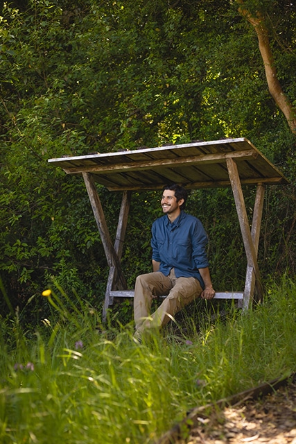 A smiling man in a blue buttondown and khaki pants sitting under a shelter enjoying in nature's beauty.